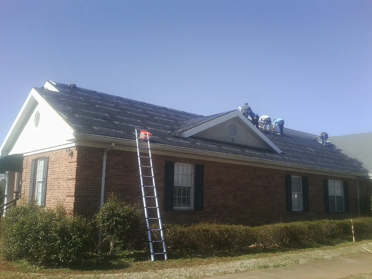 Crew tearing shingles off a brick ranch roof with a ladder set — Metal Roof Repair work by our crew in Plainville